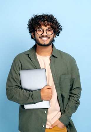 Smart handsome positive indian or arabian guy, with glasses, in casual wear, student or freelancer, holding a laptop in hand, standing on isolated blue background, looking at camera, smiling friendly